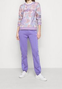 Felpa viola chiaro con motivo tie-dye e testo "CECILIE", abbinata a pantaloni della tuta viola con strisce laterali e polsini a coste.