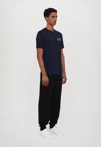 Námořnická modrá tričko s krátkým rukávem a logem, spárované s černými teplákovými kalhotami. Model obutý v bílých sportovních botách, stojí nalevo na bílém pozadí.