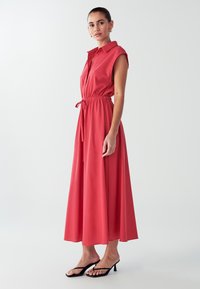 Rød, �ærmeløs skjortekjole med krave og snøre i taljen. Flydende, ankellang nederdel, parret med sorte, flade sandaler.