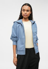 Μπλε φούτερ με φερμουάρ, με μεγάλη κουκούλα, ριγέ μανσέτες και μπροστινές τσέπες, σε συνδυασμό με ένα ανοιχτό μπεζ μπλουζάκι και μαύρο φαρδύ παντελόνι.