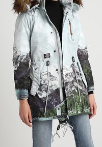 Vaaleansininen parkatakki, jossa vuoristomaisemakuosi. Takissa on irrotettava tekoturkiskaulus, kiristysnyörit vyötäröllä, vetoketjusuljenta ja vetoketjuyksityiskohtia.