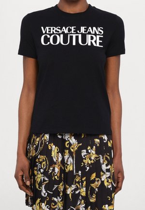 Osoba nosząca czarną koszulkę Versace Jeans Couture i plisowaną spódnicę z czarno-biało-złotym bogato zdobionym wzorem barokowym.