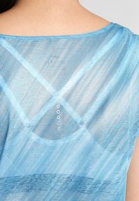 Modrý síťovaný top s texturovaným puntíkovaným vzorem. Má zaoblený výstřih a knoflíkový detail na zadní straně, který vytváří vrstvený efekt.