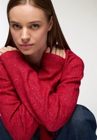 Pullover rosso lavorato a maglia con un motivo testurizzato e maniche ampie, abbinato a denim blu. La modella è seduta, abbracciando le braccia.