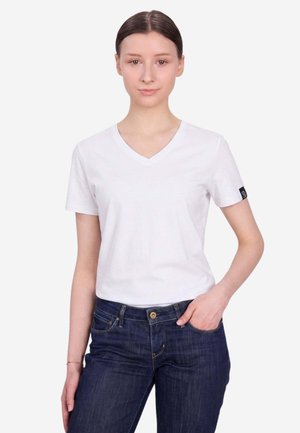 Giovane donna con capelli castani indossa una maglietta bianca con scollo a V e maniche corte e jeans blu scuro, in piedi con una mano in tasca.