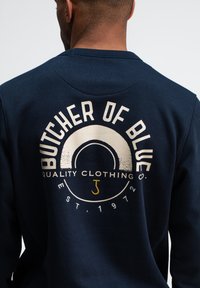 Marineblauwe sweatshirt met een circulaire grafische afbeelding op de rug met de tekst "BUTCHER OF BLUE" in wit en gele accenten; gemaakt van katoen.