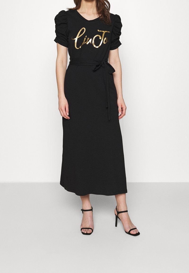 Femme portant une robe midi noire à manches courtes bouffantes, une taille nouée, des lettres dorées sur la poitrine et des talons hauts noirs à bride cheville.