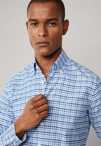 Man die de kraag van een blauw-wit geruit overhemd met knopen aanpast tegen een effen achtergrond.