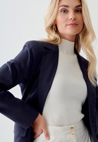 Námornícky blejzer, slim fit, štruktúrované ramená; krémový top s vysokým golierom a rebrovanou textúrou; svetlé vysoké nohavice s dvoma zlatými gombíkmi.