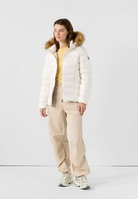 Chaqueta acolchada blanca con capucha forrada de piel, combinada con una blusa amarilla y pantalones beige. Presenta cierre con cremallera y diseño acolchado.