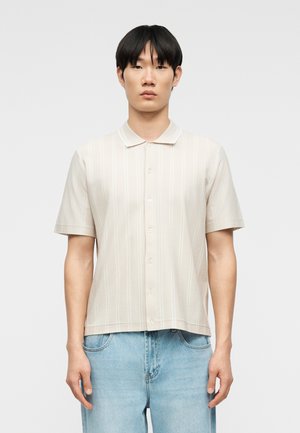 Jeune homme portant une chemise beige à manches courtes avec des rayures verticales subtiles et un jean bleu clair, debout devant un fond uni.