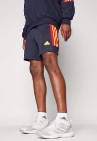 Námořnické šortky s červenými a žlutými pruhy, s vyšitým logem. Nosené s bílými sportovními botami a kotníkovými ponožkami.