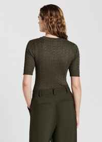 Haut en tricot à manches courtes de couleur vert olive, présentant un motif en relief de câbles, un col rond et une coupe ajustée. Associé à un pantalon taille haute.