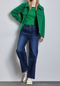 Grön texturerad jacka med två framfickor, bärs över en grön topp och mörkblå vida jeans, kombinerad med vita sneakers.