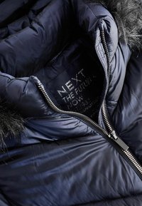 Intérieur d'une veste matelassée marine avec bord en fausse fourrure, dotée d'une fermeture éclair argentée et d'une étiquette avec le nom de la marque et les informations d'entretien. Texture lisse.