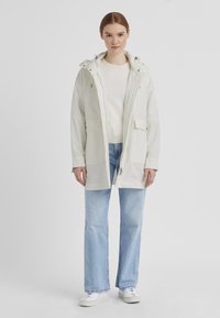 Leichte, weiße Regenjacke mit Kapuze, Reißverschluss vorne und zwei seitlichen Taschen. Getragen über einem cremefarbenen Pullover und kombiniert mit hellblauen Jeans.