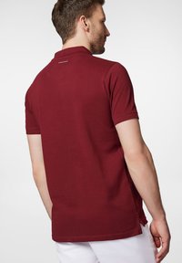 Burgundowa koszulka polo z krótkim rękawem, wykonana z bawełny. Posiada płaski kołnierzyk, boczny rozporek oraz logo w okolicy karku.