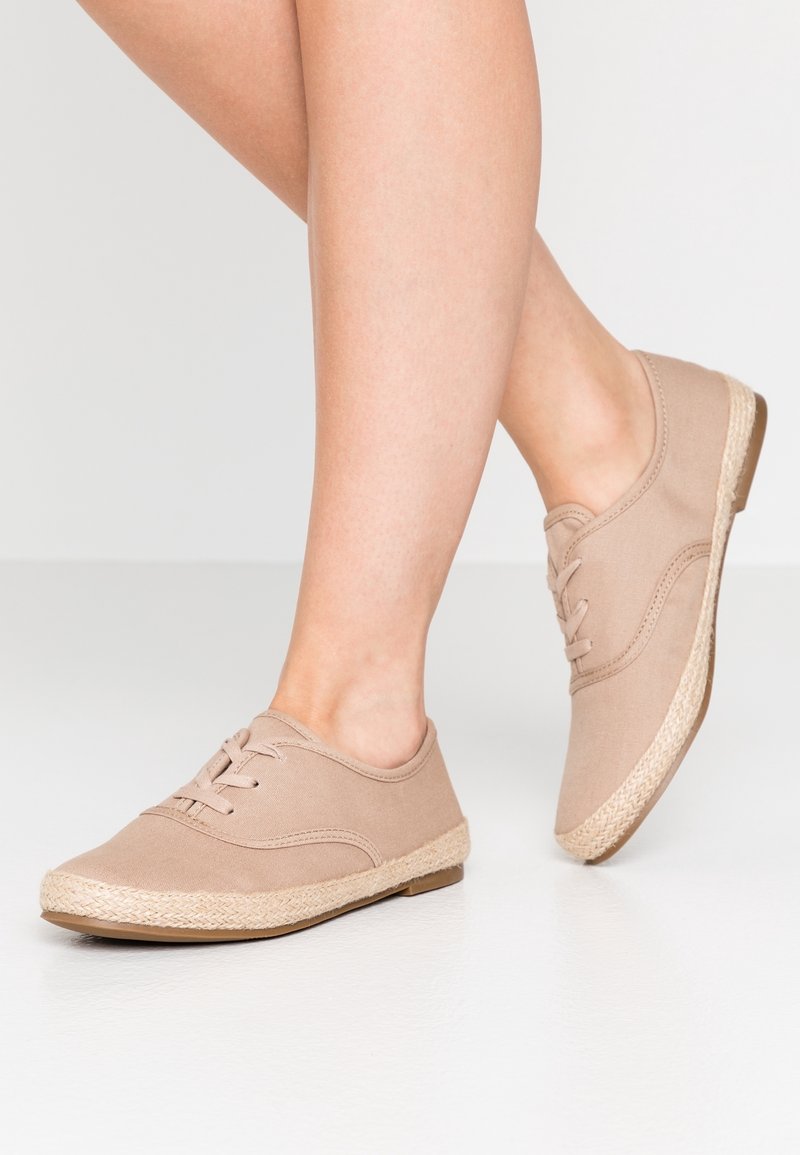 Jambes portant des chaussures décontractées en toile beige avec des lacets et un détail tissé sur la semelle, posées sur un sol blanc lisse.