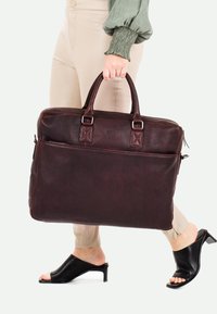 Braune Lederaktentasche mit doppelten Griffen, strukturierten Oberflächen, einer Reißverschlusstasche auf der Vorderseite und sleekem Design. Modell hält die Tasche beim Gehen.