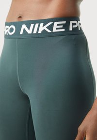 Nike Pro tajice u tamno tirkiznoj boji, s širokim crnim pojasom i bijelim brendiranjem. Tkanina je glatka, elastična i uska.