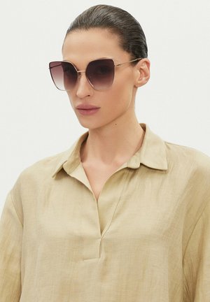 Kobieta z zaczesanymi do tyłu włosami, nosząca duże okulary przeciwsłoneczne z gradientowymi szkłami oraz beżową bluzkę z kołnierzykiem i dekoltem w serek.