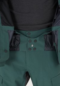 Veste imperméable verte avec accents noirs ; dotée de fermetures à pression, d'une taille élastique et d'une doublure en maille au niveau du torse.