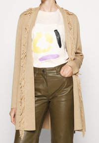 Cardigan beige avec détails à franges, superposé sur une chemise crème ornée de motifs abstraits colorés. Un pantalon en cuir vert olive complète la tenue.