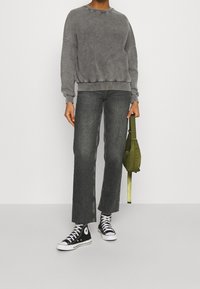 Felpa grigia sovradimensionata con orlo a coste, abbinata a jeans larghi grigio scuro e sneakers nere Converse. Tiene una piccola borsa verde.