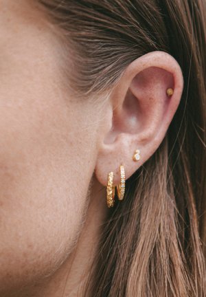 Guldøreringe med texturede overflader, der har en mindre guldstudsørering. Nærbillede af øret med lysebrunt hår i baggrunden.