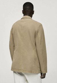 Giacca in cotone beige con un design strutturato, cuciture visibili e una vestibilità dritta. Presenta una singola cucitura centrale sul retro e un colletto standard.