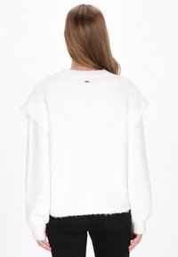 Hvid fluffy sweater med ribbet kant og skulderflapper, der har et diskret mærke bagpå. Båret med sorte tætsiddende bukser.