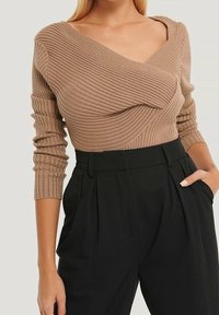 Ljusbrun ribbad stickad topp med vriden design, djup V-ringning och långa ärmar, kombinerad med svarta plisserade byxor med hög midja.