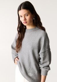 Pulover gri tricotat, cu guler înalt și canelat. Are mâneci largi și un tiv la baza cu textul "50% nu știu, 50% știu" brodat.