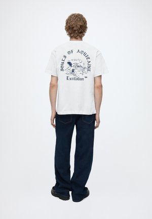 Person med krøllet hår står med ryggen til, iført en hvid grafisk t-shirt med tekst og illustration, mørke jeans og sorte sko på en ensfarvet baggrund.