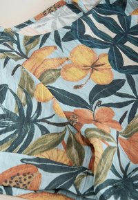 Tissu bleu clair imprimé de feuilles tropicales et de fleurs dans des tons de vert, orange et jaune, avec des détails de couture visibles.