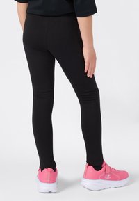 Een kind in zwarte leggings en roze sneakers, staand op een witte achtergrond, gezien van achteren.