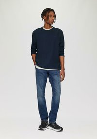 Marineblauwe geribbelde lange mouw shirt over een witte ronde hals, gecombineerd met blauwe slim-fit jeans en zwarte sportschoenen met grijze accenten.