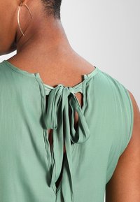 Zelený top bez rukávů s mašlí na zátylku, hladkou texturou látky a kulatým výstřihem.