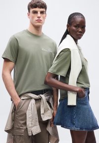 Νέος άνδρας και γυναίκα φορούν ελιάς πράσινα μπλουζάκια, με τη γυναίκα να φορά επίσης ένα λευκό πουλόβερ περασμένο στους ώμους της και μια τζιν φούστα.