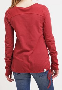 Långärmad röd topp i texturerad tyg, med rundad halsringning, justerbar dragsko längst ner och en kontrasterande ryggpaneldesign.