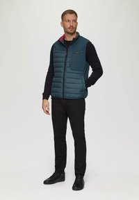 Polsterweste in Teal mit hohem Kragen, schwarzem Reißverschluss, seitlichen Taschen und einem Logoaufnäher; getragen über einem schwarzen Langarmhemd und dunklen Hosen.