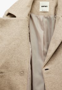 Beige wollen jas met een gladde binnenvoering. Kenmerkt zich door een genaaide kraag, knoopsluiting en zijzakken. Gelaagde buitenkant met een zachte afwerking.