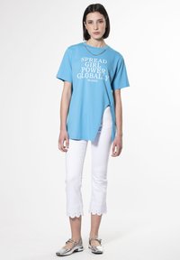 Camiseta azul cielo con texto blanco que dice "DIFUNDE EL PODER FEMENINO A NIVEL GLOBAL", combinada con pantalones blancos cortos con bordes ondulados y zapatos planos plateados.