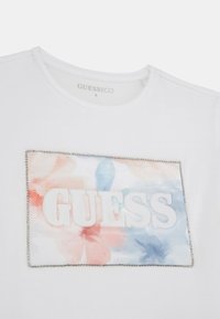 T-shirt bianco in cotone con stampa floreale ad acquerello e logo "GUESS" in rilievo, bordato da dettagli argentati.