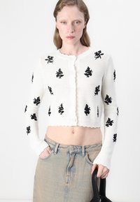 Cardigan bianco lavorato a maglia con ricami floreali neri, design cropped, bordi scallop e chiusura con bottoni abbinato a una gonna in denim a vita alta.