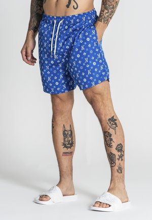 Mężczyzna noszący niebieskie szorty w wzory z białym sznurkiem, białe klapki oraz posiadający liczne czarne tatuaże na nogach i ramionach na jednolitym tle.