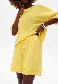 Żółty sweter dzianinowy i dopasowane szorty z fakturowanej tkaniny, luźny krój i elastyczny pas ze sznurkiem. Posiada podwinięte mankiety i minimalne detale.