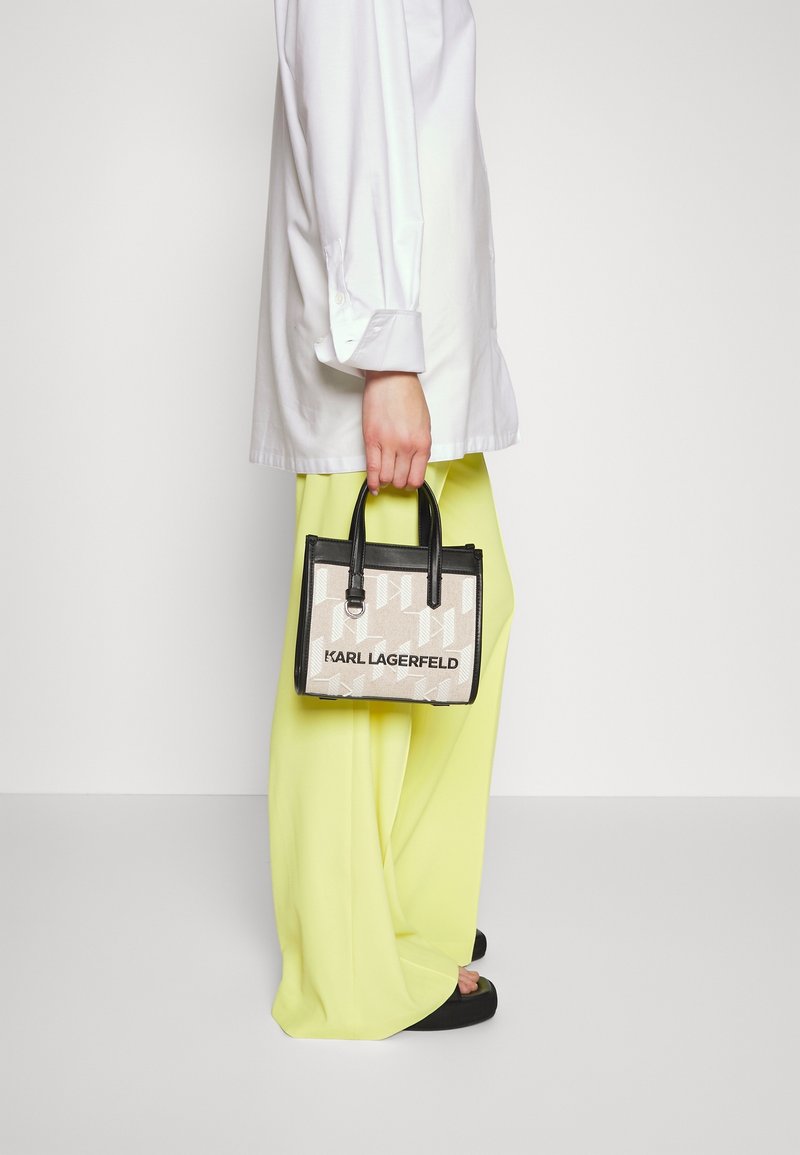 Personne portant un pantalon jaune à jambes larges et une chemise blanche, tenant un sac à main Karl Lagerfeld beige et noir par les anses.