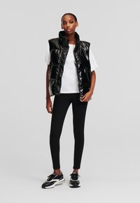 Vestă pufă neagră, lucioasă, cu guler înalt, asortată cu un tricou alb și colanți negri. Modelul poartă teniși negri.