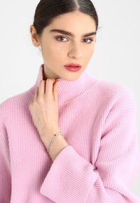 Helles rosa, geripptes Strick-Turtleneck-Oberteil mit weiten Ärmeln; Modell trägt ein silbernes Armband mit kleinen Akzenten, leicht die Hand auf das Kinn gelegt.
